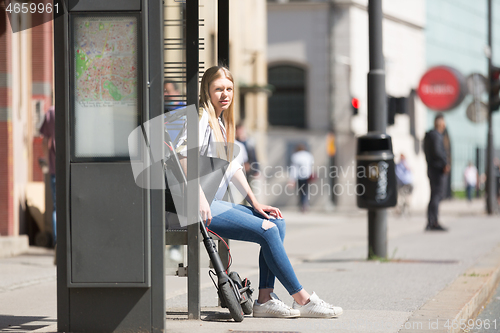 Image of Casual caucasian teenager commuter with modern foldable urban electric scooter sitting on a bus stop bench waiting for metro city bus. Urban mobility concept