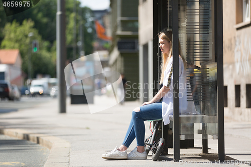 Image of Casual caucasian teenager commuter with modern foldable urban electric scooter sitting on a bus stop bench waiting for metro city bus. Urban mobility concept