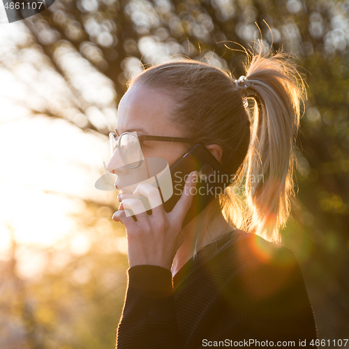 Image of Backlit rear view of young woman talking on cell phone outdoors in park at sunset. Girl holding mobile phone, using digital device, looking at setting sun