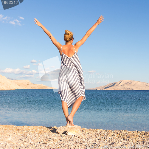 Image of Happy carefree woman rising arms, wearing beautiful striped summer dress enjoying late afternoon on white pabbled beach on Pag island, Croatia