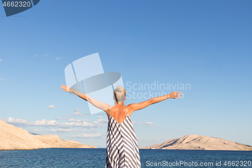 Image of Happy carefree woman rising arms, wearing beautiful striped summer dress enjoying late afternoon on white pabbled beach on Pag island, Croatia