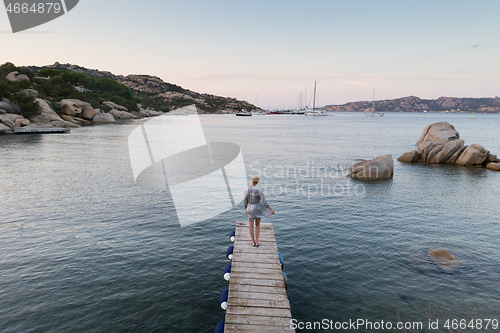 Image of Beautiful woman in luxury summer dress standing on wooden pier enjoying peaceful seascape at dusk. Vacation, resort and traveling.