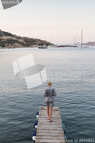 Image of Beautiful woman in luxury summer dress standing on wooden pier enjoying peaceful seascape at dusk. Vacation, resort and traveling.