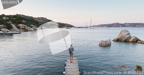 Image of Beautiful woman in luxury summer dress standing on wooden pier enjoying peaceful seascape at dusk. Vacation, resort and traveling.