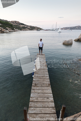 Image of Man standing on wooden pier enjoying peaceful seascape at dusk. Male tourist stands on a wooden pier in Porto Rafael, Costa Smeralda, Sardinia, Italy