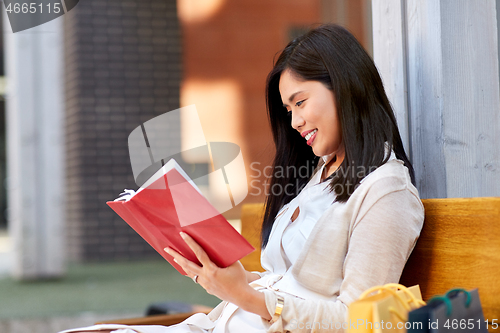 Image of smiling asian woman reading book sitting on bench
