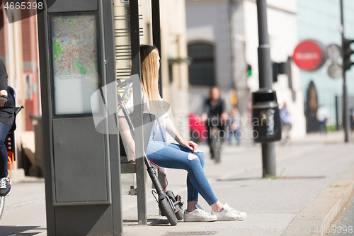 Image of Casual caucasian teenager commuter with modern foldable urban electric scooter sitting on a bus stop bench waiting for metro city bus. Urban mobility concept