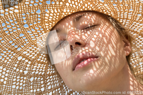 Image of Close-up portrait of woman, eyes shut, wearing straw hat enjoying summer sun. Pattern of shadows falling on her face