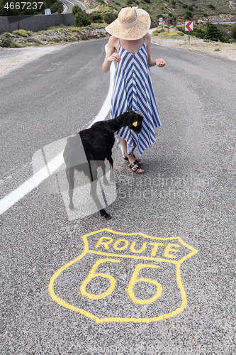 Image of Young attractive woman wearing striped summer dress and straw hat standing on an endless straight empty road in the middle of nowhere on the Route 66 road and feeding black sheep.