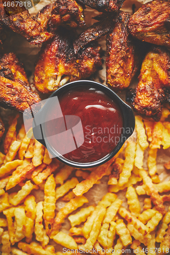 Image of Appetizing roasted chicken wings and french fries with barbecue dip, served on baking paper