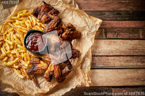 Image of Appetizing roasted chicken wings and french fries with barbecue dip, served on baking paper