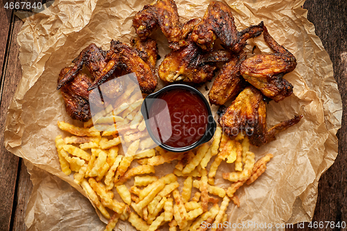 Image of Appetizing roasted chicken wings and french fries with barbecue dip, served on baking paper