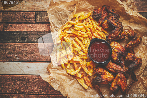 Image of Appetizing roasted chicken wings and french fries with barbecue dip, served on baking paper