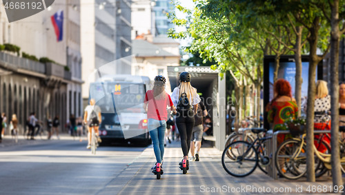 Image of Rear view of trendy fashinable teenager girls riding public rental electric scooters in urban city environment. New eco-friendly modern public city transport in Ljubljana, Slovenia