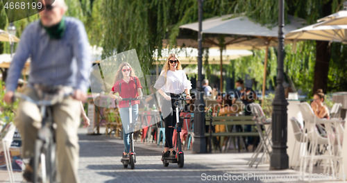 Image of Trendy fashinable teenager girls riding public rental electric scooters in urban city environment. New eco-friendly modern public city transport in Ljubljana, Slovenia