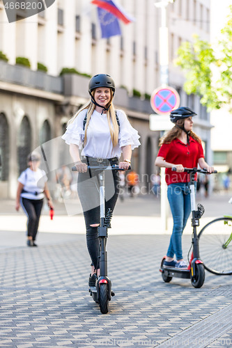 Image of Trendy fashinable teenager girls riding public rental electric scooters in urban city environment. New eco-friendly modern public city transport in Ljubljana, Slovenia