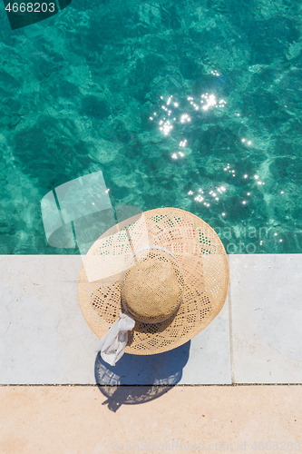 Image of Woman wearing big summer sun hat relaxing on pier by clear turquoise sea.