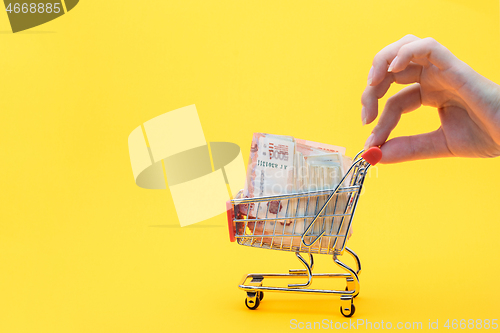 Image of Hand holds the handle of a grocery cart with a pack of five thousandth Russian rubles, yellow background