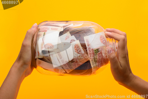 Image of Hands holding a three-liter jar in which lie five thousandth bills, yellow background