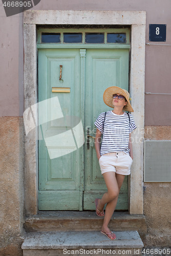 Image of Beautiful young female tourist woman wearing sun hat, standing and relaxing in shade in front of vinatage wooden door in old Mediterranean town while sightseeing on hot summer day