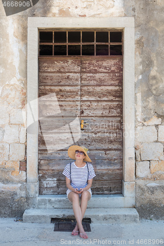 Image of Beautiful young female tourist woman sitting and resting on vinatage wooden doorstep and textured stone wall at old Mediterranean town, smiling, holding, using smart phone to network on vacationes