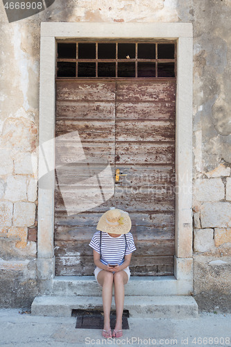 Image of Beautiful young female tourist woman sitting and resting on vinatage wooden doorstep and textured stone wall at old Mediterranean town, smiling, holding, using smart phone to network on vacationes