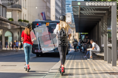 Image of Rear view of trendy fashinable teenager girls riding public rental electric scooters in urban city environment. New eco-friendly modern public city transport in Ljubljana, Slovenia