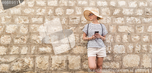 Image of Beautiful young female tourist woman standing in front of old textured stone wall at old Mediterranean town, smiling, holding, smart phone to network on vacationes. Copy space