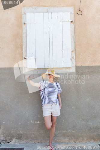 Image of Beautiful young female tourist woman wearing big straw hat, taking self portrait selfie, standing in front of white vinatage wooden window and textured stone wall at old Mediterranean town