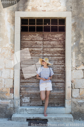 Image of Beautiful young female tourist woman standing in front of vinatage wooden door and textured stone wall at old Mediterranean town, smiling, holding, smart phone to network on vacationes