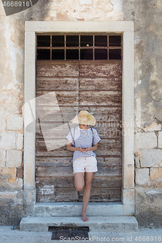 Image of Beautiful young female tourist woman standing in front of vinatage wooden window and textured stone wall at old Mediterranean town, smiling, holding, using smart phone to network on vacationes