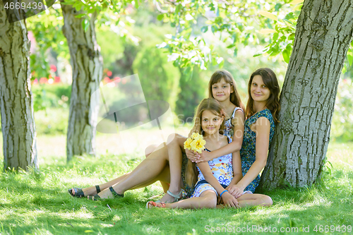 Image of Mom with two daughters sits on green grass near tree in park