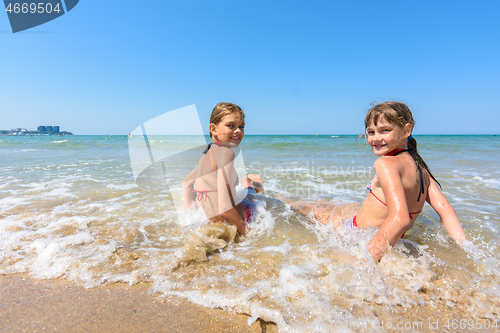 Image of Two girls sit on the sea coast and turn around happily looking at the frame