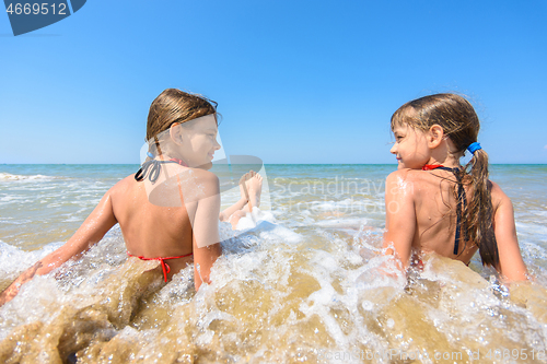 Image of Children sit in water on a sandy beach and look at each other happily