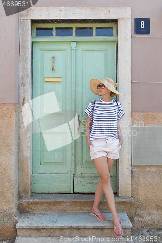 Image of Beautiful young female tourist woman wearing sun hat, standing and relaxing in shade in front of vinatage wooden door in old Mediterranean town while sightseeing on hot summer day