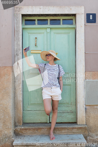 Image of Beautiful young female tourist woman wearing big straw hat, taking self portrait selfie, standing in front of turquoise vinatage wooden door and textured stone wall at old Mediterranean town