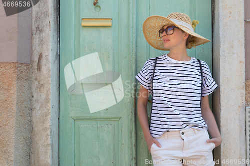 Image of Beautiful young female tourist woman wearing sun hat, standing and relaxing in shade in front of vinatage wooden door in old Mediterranean town while sightseeing on hot summer day