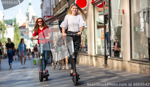 Image of Trendy fashinable teenager girls riding public rental electric scooters in urban city environment. New eco-friendly modern public city transport in Ljubljana, Slovenia