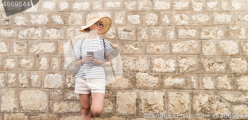 Image of Beautiful young female tourist woman standing in front of old textured stone wall at old Mediterranean town, smiling, holding, smart phone to network on vacationes. Copy space