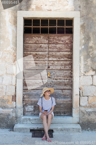 Image of Beautiful young female tourist woman sitting and resting on vinatage wooden doorstep and textured stone wall at old Mediterranean town, smiling, holding, using smart phone to network on vacationes