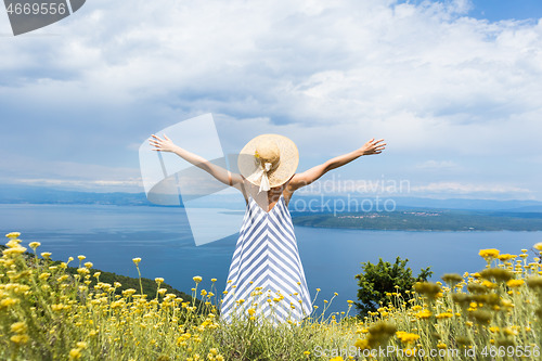 Image of Rear view of joung woman wearing striped summer dress and straw hat standing in super bloom of wildflowers, relaxing with hands up to the sky, enjoing beautiful view of Adriatic sea nature, Croatia