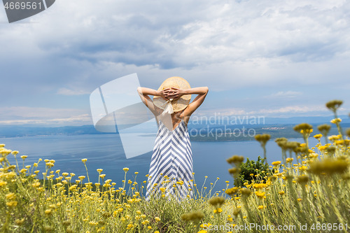Image of Rear view of young woman wearing striped summer dress and straw hat standing in super bloom of wildflowers, relaxing while enjoing beautiful view of Adriatic sea nature, Croatia