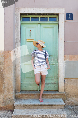 Image of Beautiful young female tourist woman wearing sun hat, standing and relaxing in shade in front of vinatage wooden door in old Mediterranean town while sightseeing on hot summer day