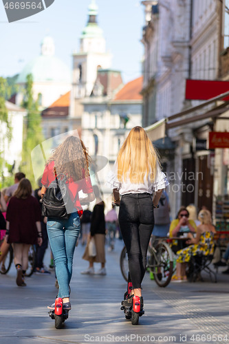 Image of Rear view of trendy fashinable teenager girls riding public rental electric scooters in urban city environment. New eco-friendly modern public city transport in Ljubljana, Slovenia