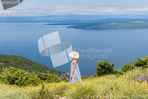 Image of Rear view of young woman wearing striped summer dress and straw hat standing in super bloom of wildflowers, relaxing while enjoing beautiful view of Adriatic sea nature, Croatia