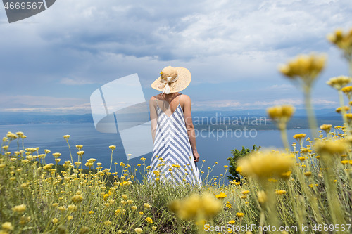 Image of Rear view of young woman wearing striped summer dress and straw hat standing in super bloom of wildflowers, relaxing while enjoing beautiful view of Adriatic sea nature, Croatia