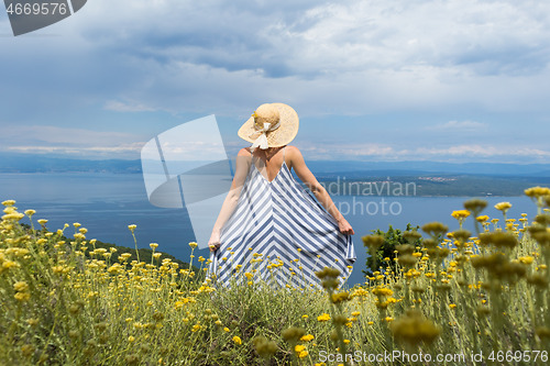 Image of Rear view of young woman wearing striped summer dress and straw hat standing in super bloom of wildflowers, relaxing while enjoing beautiful view of Adriatic sea nature, Croatia