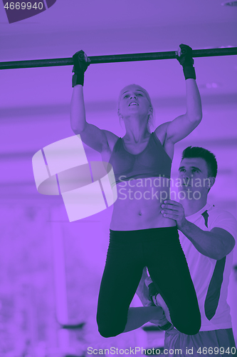 Image of trainer support young woman while lifting on bar in fitness gym