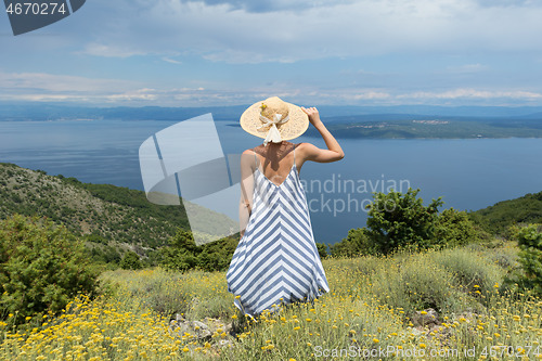 Image of Rear view of young woman wearing striped summer dress and straw hat standing in super bloom of wildflowers, relaxing with hands up to the sky, enjoing beautiful view of Adriatic sea nature, Croatia