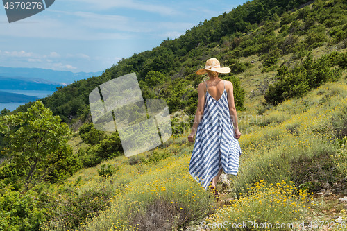 Image of Young woman wearing striped summer dress and straw hat walking among super bloom of wildflowers, relaxing while enjoing beautiful nature of of Adriatic sea coastal nature of Croatia